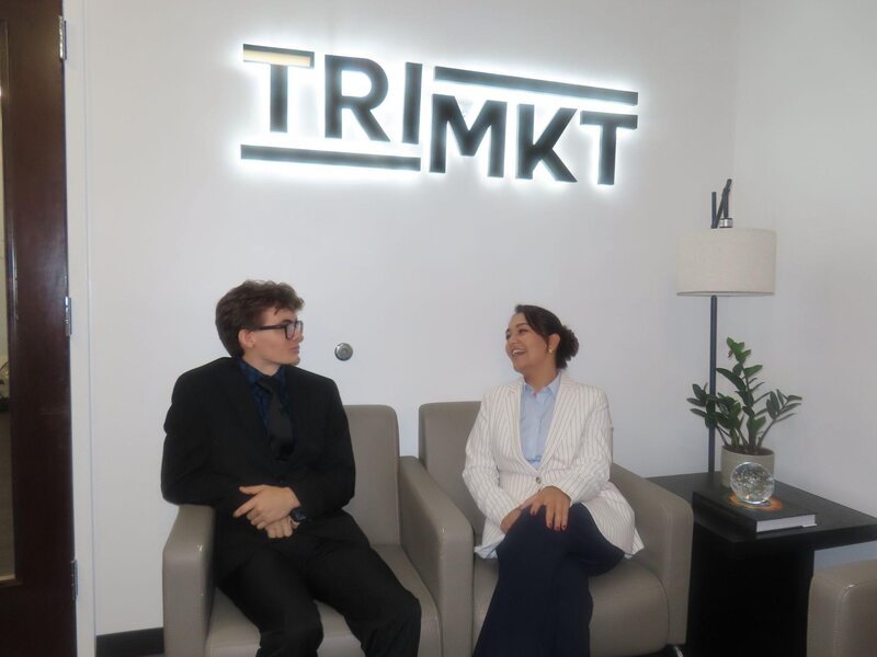 Two TriMkt team members talking beneath the TriMkt sign in the Raleigh office